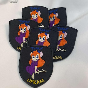 May include: Embroidered black patches featuring a cartoon character with orange hair, a purple dress, and the word "Tайка" in blue. The patches are shield-shaped and have white stitching. Some patches have additional text.