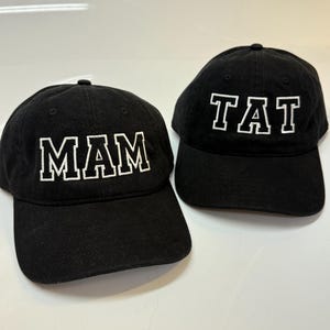 May include: Two black baseball caps with white embroidered lettering. One cap features the text MAM in a bold collegiate font, while the other cap displays the text TAT in the same white embroidered style. Both caps have a curved brim.