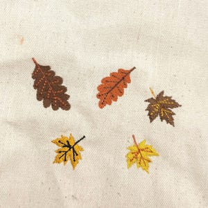 May include: Close-up of a cream-colored fabric with embroidered autumn leaves. The leaves are in shades of brown, orange, and yellow, with detailed stitching. The design evokes a fall or harvest theme.