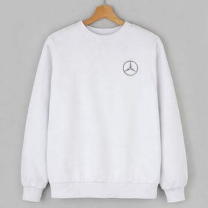 Può includere: Felpa bianca girocollo con un piccolo logo Mercedes-Benz grigio sul petto sinistro. La felpa ha maniche lunghe e polsini e orlo a coste. Appesa a una gruccia di legno su uno sfondo grigio.