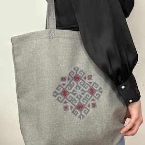 Pode incluir: Uma bolsa tote cinza com um design geométrico bordado em vermelho e azul. A bolsa é feita de um material semelhante ao linho.