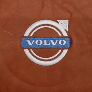 May include: A close-up shot of a brown leather surface with a Volvo logo embroidered on it. The logo consists of a white circle, a blue rectangle with the word "VOLVO" in white, and a classic design.