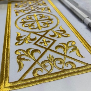 Op de afbeelding: Detailopname van witte stof met ingewikkelde gouden borduursels. Het ontwerp toont een centraal kruis in een cirkel, omringd door geometrische vormen en bloemmotieven. De borduursels zijn gedetailleerd en lijken machinaal te zijn gemaakt.