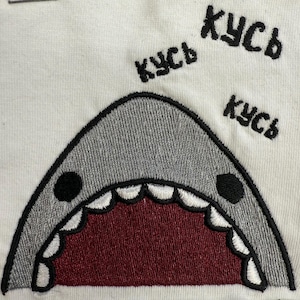 Può includere: Tessuto bianco con un ricamo a forma di bocca di squalo. La bocca dello squalo è aperta, rivelando un interno rosso e denti bianchi. Sopra lo squalo, la parola "кусь" è ricamata tre volte in nero.
