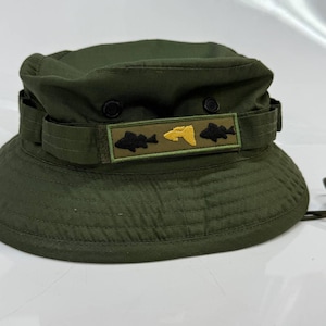 Puede incluir: Sombrero boonie verde oliva con ala ancha y barboquejo. El sombrero presenta un parche rectangular con peces bordados y un pez amarillo en el centro. El sombrero está hecho de tela duradera.