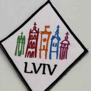 Puede incluir: Parche bordado en forma de diamante con fondo blanco y borde negro. Presenta un colorido paisaje urbano y la palabra "LVIV" en negro. El paisaje urbano incluye edificios en verde, rojo, naranja, azul y morado.