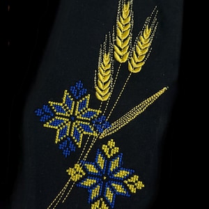 Puede incluir: Tela negra con bordados de espigas de trigo y diseños florales en forma de estrella en amarillo y azul. El bordado tiene una apariencia detallada y texturizada, creando un contraste llamativo sobre el fondo oscuro.