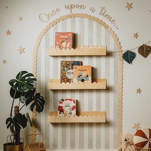 Op de afbeelding: Een grillige kinderkamerscène met houten planken met kinderboeken. Een bruine berenuitsparing houdt een luchtballonlamp vast. Decoratieve sterren en een bladguirlande sieren de muur. Een teddybeer en een rieten mand dragen bij aan de gezellige sfeer.