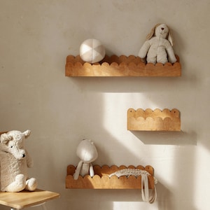 Peut inclure: Trois étagères en bois avec un bord festonné sont montées sur un mur de couleur claire. Les étagères contiennent des animaux en peluche, dont un ours, un lapin et un hippopotame. Un jouet teckel en bois est posé sur le sol.