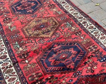 May include: A red, blue, and cream colored vintage rug with a geometric pattern. The rug has a fringe border and is made of wool.