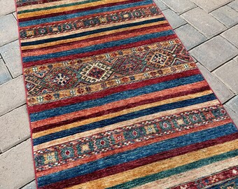 Könnte beinhalten: Ein langer, schmaler Teppich mit einem farbenfrohen, geometrischen Muster. Der Teppich hat Streifen in Rot, Blau, Gelb und Grün und zeigt im Zentrum komplizierte Designs.