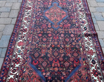 May include: A vintage Persian rug with a dark red background and intricate floral and geometric patterns in shades of blue, red, and white. The rug has a white border with a repeating design.