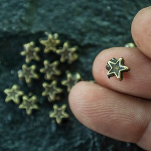 May include: A close-up of a hand holding a small, gold-colored star-shaped metal charm. The charm has a slightly raised, black star design in the center. There are several other similar charms scattered on a dark green surface.