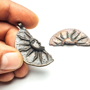 Puede incluir: Un colgante de bronce en forma de sol con una cara. El colgante está roto en dos piezas, una pieza está en una mano y la otra está sobre una superficie blanca.