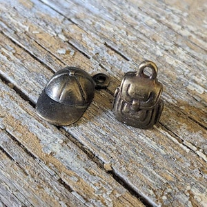 May include: Two small, gold-coloured metal charms. One charm is shaped like a baseball cap and the other is shaped like a backpack.