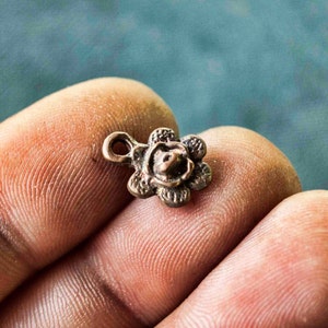 May include: A small, bronze-coloured flower charm with a loop for attaching to a necklace or bracelet. The charm has a detailed design with a raised centre and five petals.
