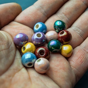 Può includere: Una collezione di perline in ceramica colorate in varie tonalità di blu, verde, giallo, viola e marrone. Le perline sono lisce e lucide, con un leggero bagliore iridescente.
