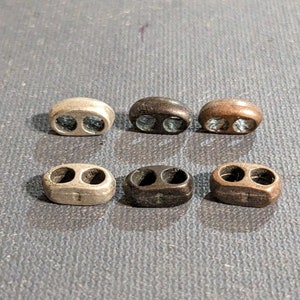 Könnte beinhalten: Sechs ovale Kordelstopper in verschiedenen Metallausführungen: Silber, Schwarz und Bronze. Jeder Kordelstopper hat zwei Löcher. Die Kordelstopper sind auf einer grauen Oberfläche angeordnet.