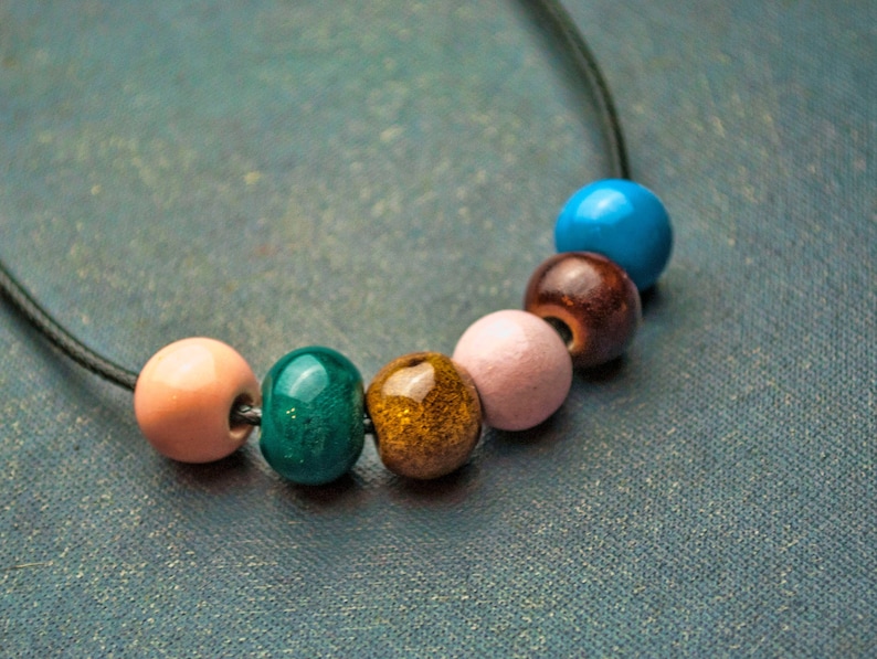 May include: A necklace with six colourful beads, including pink, green, brown, blue, and gold. The beads are strung on a black cord.