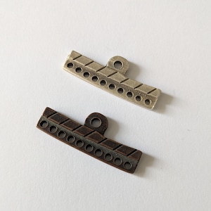 Op de afbeelding: Twee metalen schuifgespen, een zilveren en een bronzen, met een decoratief patroon en een lus aan de bovenkant.