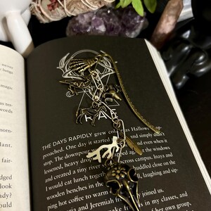 Puede incluir: Un marcapáginas de bronce con varios amuletos, incluyendo una calavera de cuervo, una mano de esqueleto y una silueta de ciervo, descansa sobre un libro abierto. El marcapáginas tiene un diseño largo y delgado y está hecho de metal. Las páginas del libro son visibles, con texto impreso.