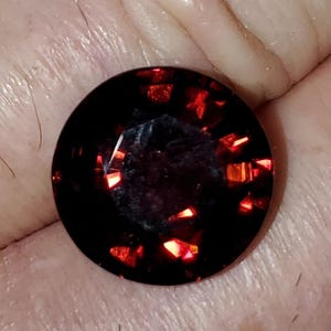 May include: A faceted, round, dark red gemstone with bright red internal reflections. The gem is set against a neutral background, showcasing its deep color and brilliance. The cut of the stone creates multiple facets that catch and reflect light.