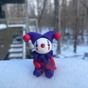 May include: A small, purple and red jester figurine with a white face and red painted lips and cheeks. The figurine is sitting in the snow.