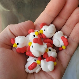May include: A hand holding a group of small, white, clay chickens with red and yellow accents. The chickens have small, black eyes and are all facing forward.