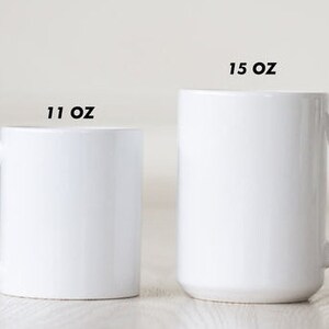 May include: Two white ceramic mugs, one labeled "11 OZ" and the other "15 OZ". The mugs have a simple, cylindrical design with a curved handle. The mugs are displayed on a light-colored surface.