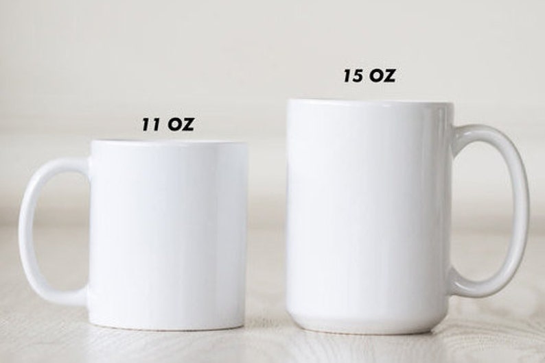 May include: Two white ceramic mugs, one labeled "11 OZ" and the other "15 OZ". The mugs have a simple, cylindrical design with a curved handle. The mugs are displayed on a light-colored surface.
