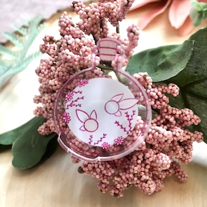 May include: A clear glass bottle charm with a pink outline and a white background. The bottle has a pink floral design and two pink bunnies inside.
