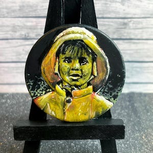 Puede incluir: Un retrato redondo pintado a mano de una persona con un abrigo con capucha amarillo sobre un fondo negro. La obra de arte se exhibe en un pequeño caballete negro. La cara de la persona está pintada con tonos verdes y amarillos.