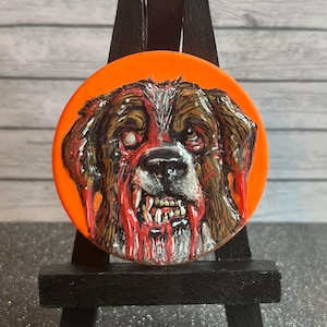 Puede incluir: Un diseño redondo de perro zombi con fondo naranja. El perro tiene pelaje marrón y blanco, con sangre roja goteando de su boca y ojo. El diseño se muestra en un pequeño caballete negro.