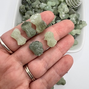 May include: A bowl filled with small, green, and white, translucent, crystal-like stones. A hand is holding a few of the stones, which are shaped like small, irregular, geometric forms.