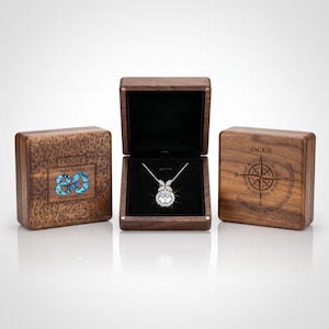 May include: A silver-toned necklace with a round pendant and leaf-shaped accents inside an open wooden box. Two closed wooden boxes are present, one engraved with "DENISE Happy Birthday 80 29TH SEPTEMBER 2026" and the other with "JACKIE" and a compass design.