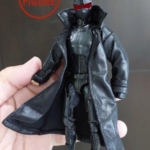 以下が含まれることがあります： アクションフィギュア用の黒いテクスチャード加工のトレンチコート。幅広の襟と長袖が特徴です。このコートは、赤いバイザーのディテールがある黒いスーツの上に着用するように設計されています。左上の赤い円形のスタンプには「NO FIGURE」と書かれています。