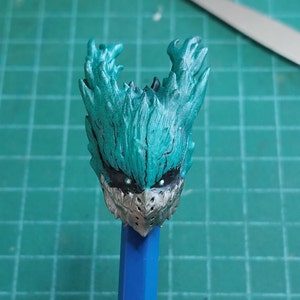 Pu&ograve; includere: Una testa di figura in plastica verde e grigia con un design dettagliato. La testa della figura ha una parte superiore appuntita e un viso a forma di maschera con due occhi neri.