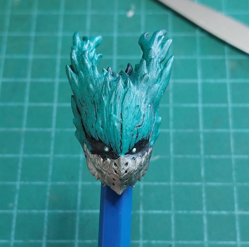 Pu&ograve; includere: Una testa di figura in plastica verde e grigia con un design dettagliato. La testa della figura ha una parte superiore appuntita e un viso a forma di maschera con due occhi neri.