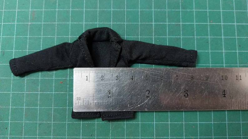 Op de afbeelding: Zwart langarm shirt met een kraag, plat gelegd op een groene ondergrond met een liniaal die 17,8 cm lang is.