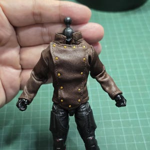 Puede incluir: Una chaqueta marrón en miniatura con botones dorados. La chaqueta es parte de una pequeña figura de acción, con pantalones y guantes negros. La cabeza de la figura es negra y tiene una rótula.