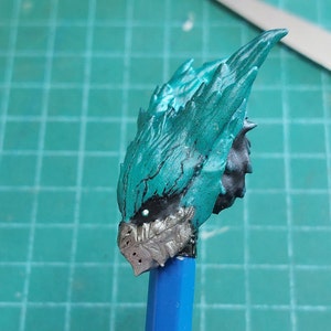 Pu&ograve; includere: Primo piano di una testa di modello dipinta in turchese e argento con un design a punta e texture. La testa &egrave; montata su una matita blu.