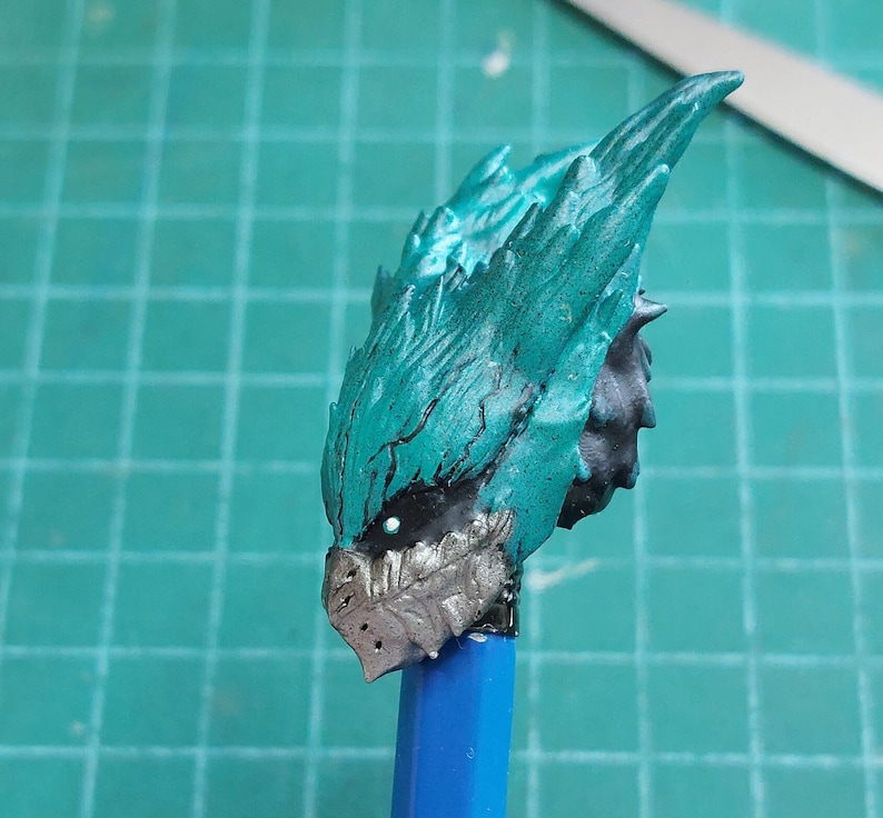 Pu&ograve; includere: Primo piano di una testa di modello dipinta in turchese e argento con un design a punta e texture. La testa &egrave; montata su una matita blu.