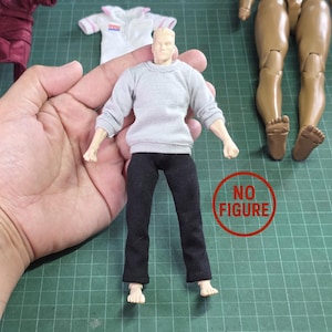 May include: A doll figure wearing a gray sweatshirt and black pants. The doll has a fair complexion and short, blond hair. Other doll clothing items are visible in the background.