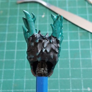 Pu&ograve; includere: Una figura in miniatura nera e turchese con una testa spinosa e frondosa. La figura &egrave; montata su una matita blu.