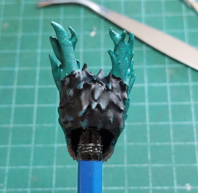 Pu&ograve; includere: Una figura in miniatura nera e turchese con una testa spinosa e frondosa. La figura &egrave; montata su una matita blu.