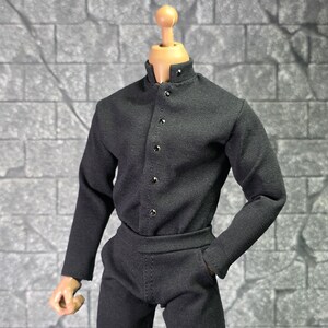 Puede incluir: Conjunto negro para muñeca, que incluye una camisa de manga larga con cuello mao y cierre de botones, y pantalones a juego. El conjunto se muestra sobre un cuerpo de muñeca con tono de piel neutro. La tela parece ser lisa y mate.