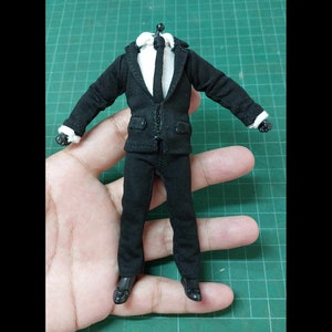 Op de afbeelding: Een zwart pak met een wit overhemd en een zwarte stropdas. Het pak is gemaakt voor een action figure.