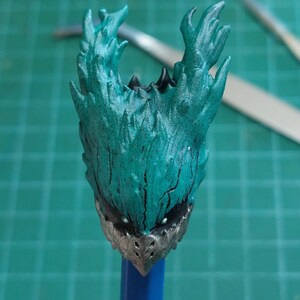 Pu&ograve; includere: Una testa di action figure verde acqua e argento con un design a forma di fiamma spinosa. La testa &egrave; montata su una matita blu.