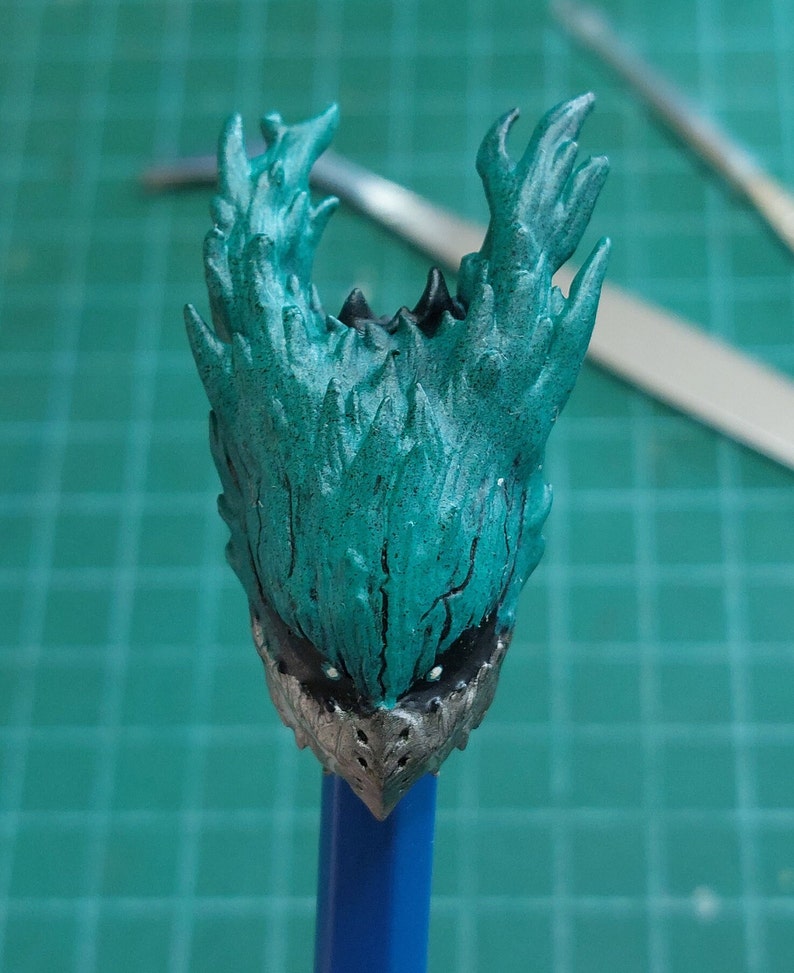 Pu&ograve; includere: Una testa di action figure verde acqua e argento con un design a forma di fiamma spinosa. La testa &egrave; montata su una matita blu.