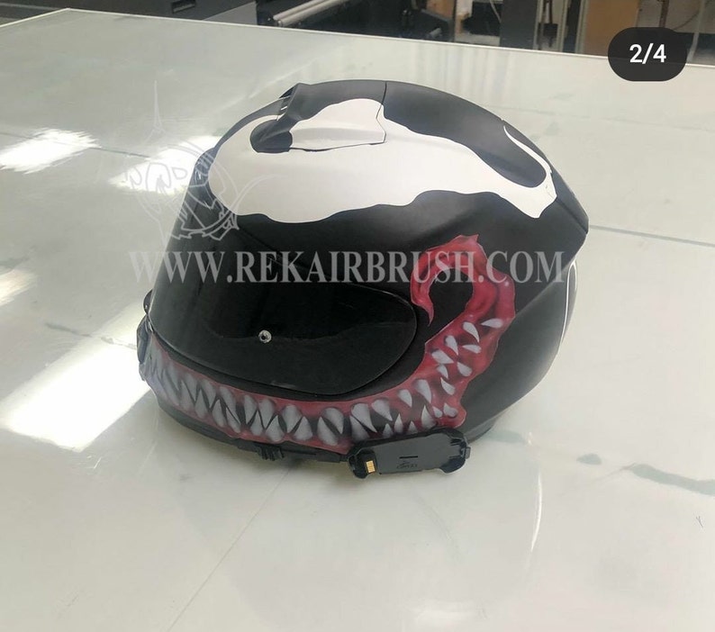 Puede incluir: Casco de moto negro con dise&ntilde;o Venom personalizado. El casco presenta detalles en blanco y rojo, incluyendo un dise&ntilde;o detallado de boca y dientes. La direcci&oacute;n del sitio web www.rekairbrush.com es visible.